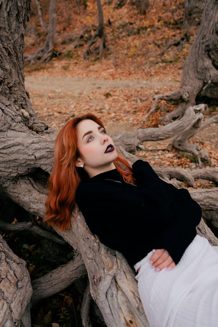 Minust A woman with red hair relaxes against tree roots in a serene autumn setting.